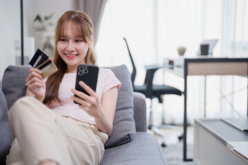 Happy Asian woman relaxing on a comfortable sofa, using her smartphone and credit card for online...