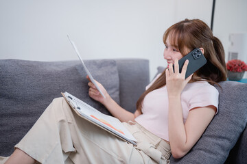 Young asian businesswoman working from home sofa talking on smartphone and analyzing financial...