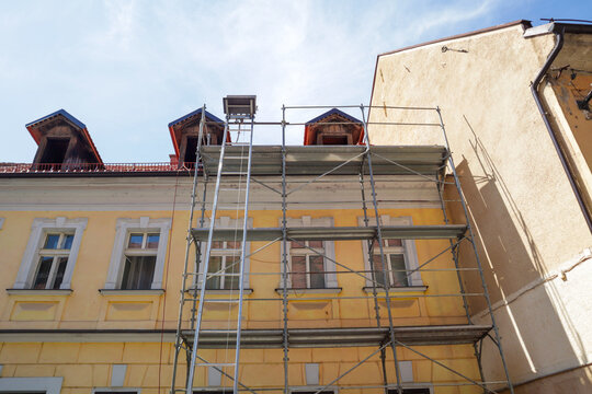 Scaffolding is erected along the facade of a light yellow building with multiple windows and red roof details, indicating construction or renovation work.
