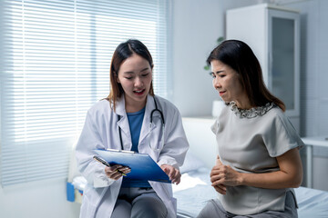 A woman is sitting on a hospital bed while a doctor looks at her medical records