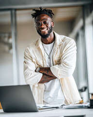 A confident young man with glasses and tattoos smiles warmly while standing with crossed arms in a modern workspace, wearing a casual white shirt.