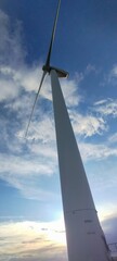 A tall wind turbine tower is seen against a blue sky. The sky is partly cloudy, but the sun is shining through the clouds. Concept of energy and progress
