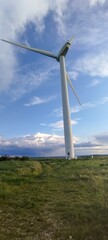 A wind turbine is standing tall in a field. The sky is clear and blue, with a few clouds scattered throughout. Concept of calm and serenity