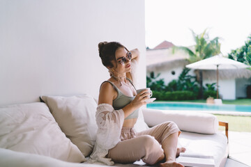 Young freelancer woman holding coffee, slightly tilting head in thoughtful pause. Image reflects...