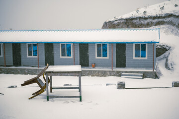 Wooden cabins in a snowy mountain region. An overturned plastic chair sits on a wooden table covered in fresh snow. Winter landscape with falling snow and overcast sky.