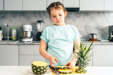 Happy little girl cutting pineapple on the background of the kitchen at home. Pre-teen girl smiling while preparing healthy fruits for eating. Selected focus.
