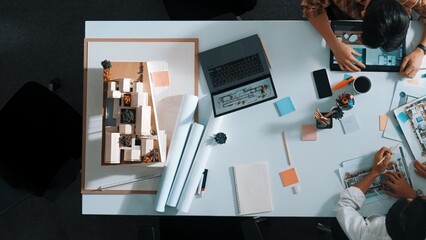 Top aerial view business people brainstorming about building interior at table with house model and laptop displayed house plan while architect engineer drawing and writing at blueprint. Alimentation.