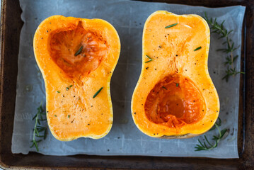Seasoned butternut squash halves on baking tray with herbs.