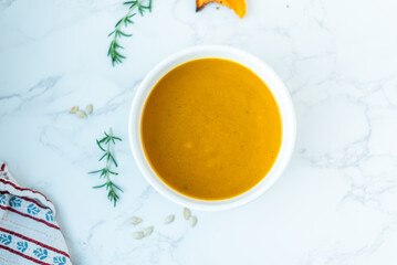 A smooth bowl of butternut squash soup sits on a white marble surface, capturing a light and...