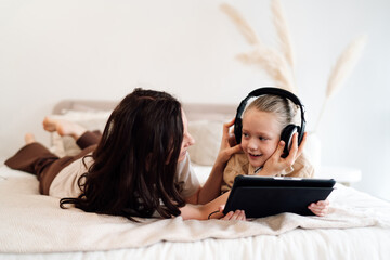 Young woman helps smiling daughter adjust headphones while they lie on bed with tablet,...