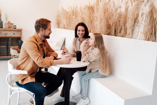 Father relaxes with coffee as mother and daughter share moment, illustrating role shifts, digital breaks, and calm modern fatherhood inside a serene home-inspired cafe.