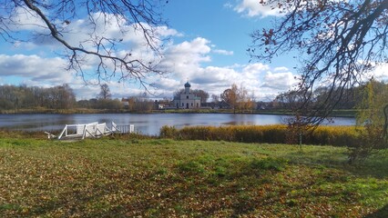 Oak trees grow on the grassy shore of the lake. The leaves crumbled. Reeds grow in the water near the wooden pier. On the far shore stands an Orthodox church with a dome and a cross. Sunny autumn