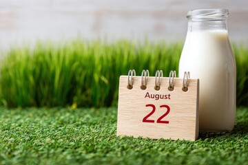 August 22 date on a wooden calendar stands alongside a milk bottle set on green turf with a backdrop of tall grass