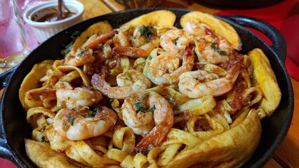 Close-up of a delicious shrimp and pasta dish with fried plantains served in a rustic black cast-iron skillet on a wooden table. A traditional seafood meal with fresh herbs and fettuccine.