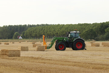 a tractor with a front loader is loading big straw bales in field in the dutch countryside in summer