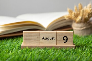 Wooden calendar blocks show August 9 on grass beside an open book  wheat bundle