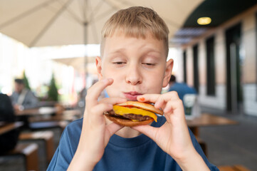 Teen blond boy at summer cafe with burger in hand, relaxing outdoors
