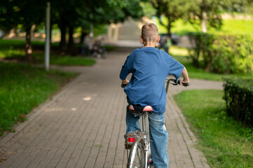 Blond teenage boy riding bicycle through park, rear view on sunny summer day