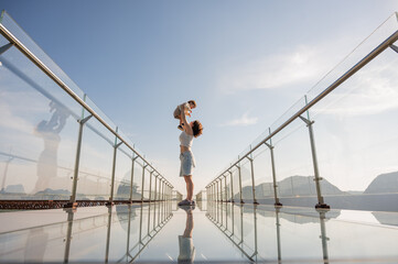 Caucasian woman holds her little son in her arms while standing on a glass bridge in the mountains. 