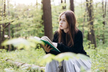 Teenage girl reading a book while sitting on a log in the forest, copy space.