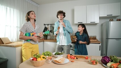 Caucasian skilled father, mother and asian daughter making breakfast while dancing together. Skilled mom playing ukulele while preparing vegetable at modern kitchen. Healthy food concept. Pedagogy.