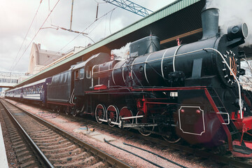 A classic black steam train with red accents emits smoke while stopped at a railway station platform, evoking vintage travel nostalgia