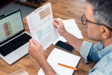 Grey haired tenant man holds printed utility bill at table with paper notebook and laptop, male...