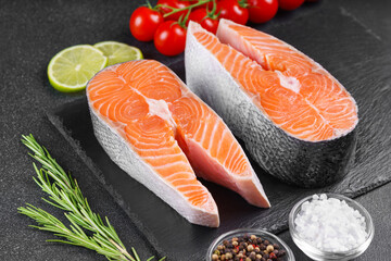Two fresh raw salmon or trout steaks on black stone surface with food ingredients. Tomatoes, lime, rosemary, salt, and pepper. Healthy eating, seafood, cooking, recipe and menu concepts.