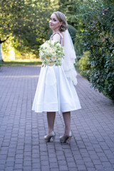 Stylish bride in a tea-length white wedding dress and lace veil, holding a bouquet of ivory and...