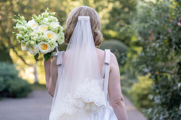 Back view of a bride in a white wedding dress with a long veil, holding an elegant bouquet of ivory...