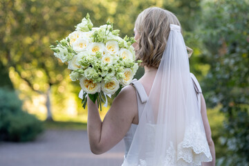 Back view of a bride in a white wedding dress with a long veil, holding an elegant bouquet of ivory...