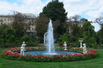 circular garden fountain with cherub statues and vibrant flowers set in a city park, against a...