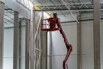 Worker on red articulated boom lift installing or inspecting ceiling structure inside industrial warehouse or factory construction site with high concrete walls and steel beams