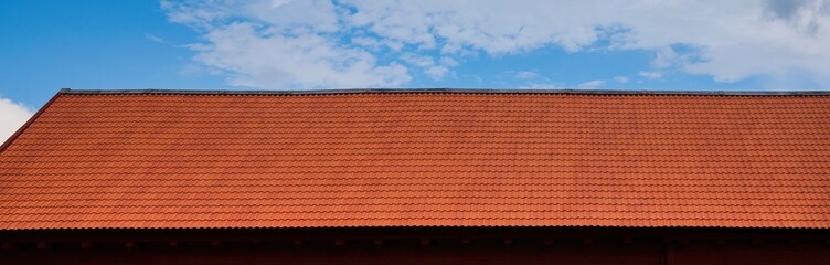 Red Tiled Roof Against Blue Sky. Panoramic view of a traditional red clay tiled roof with a clear blue sky and scattered clouds in the background.