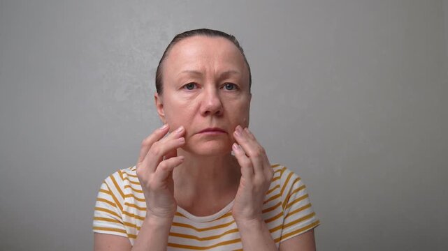 50-55-year-old woman looks with chagrin at her face with wrinkles, puffiness, and flabby skin. Age-related changes and fatigue. A woman is planning anti-aging procedures or plastic surgery