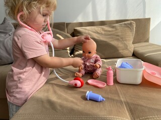 Little girl listens to a doll’s chest using a toy stethoscope. Playing doctor to learn care and...