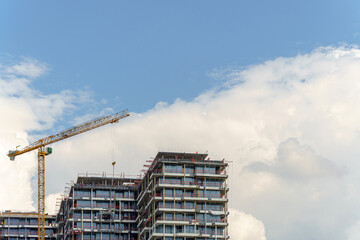 Construction of a multi-unit residential building is underway with the help of a tower crane against a backdrop of blue sky and clouds. 