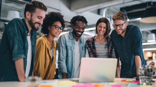 A group of smiling, diverse young professionals enthusiastically collaborate and brainstorm, gathered around a laptop in a bright, contemporary office environment