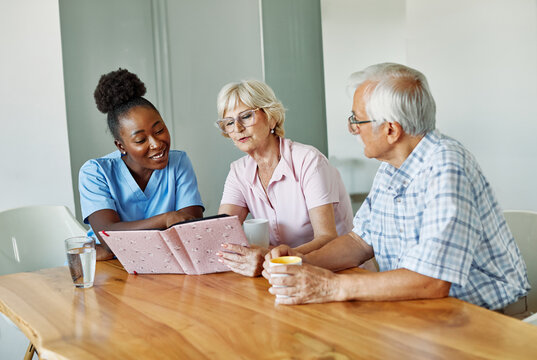 Doctor or nurse caregiver with senior woman and man, a senior couiple, looking at photo album and picture or book or brochure at home or nursing home - Powered by Adobe