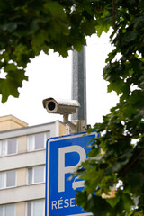 Surveillance camera watches silence. Parking sign beneath it whispers order. Apartment windows stay...