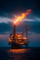 Obraz premium An oil rig stands tall in the ocean as flames erupt from its top, illuminating the dark sky. The blue water reflects the fiery glow amid a moody, cloudy backdrop
