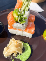 Sushi dish featuring stacked salmon rolls garnished with fresh vegetables, accompanied by wasabi and pickled ginger on a dark plate, showcasing vibrant colors and textures