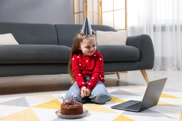 Birthday celebration. Little girl with cake having video call via laptop at home