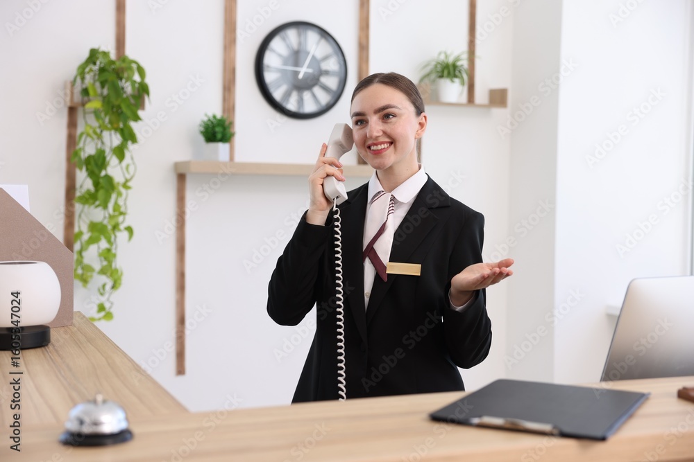 Poster smiling receptionist talking with client by phone at reception desk in hotel