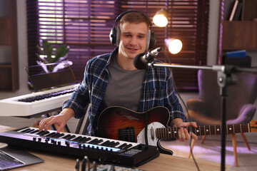 Smiling man in headphones working with MIDI keyboard and microphone at table in home studio