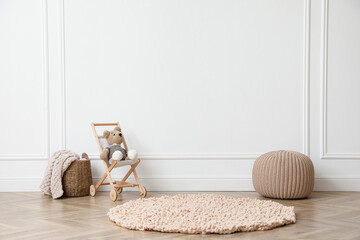 Toy stroller with bear, pouf and wicker basket near white wall in child room. Interior design