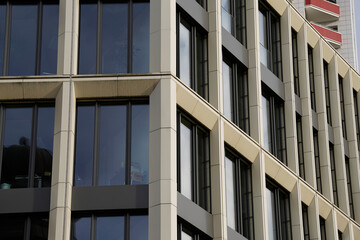 Modern office building with beige vertical panels and large dark-framed windows. Interior furniture visible through clear glass shows minimalist and professional design.

