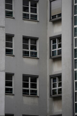 Close-up of light gray building facade with several double-pane windows featuring white frames. The texture of the wall contrasts with smooth glass surfaces, and nearby green foliage adds natural soft