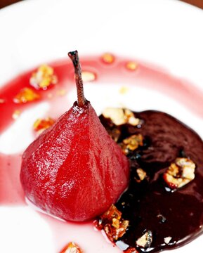 A luxurious close-up of a red wine-poached pear plated with a dark berry reduction, chocolate sauce,  and crunchy nut brittle. 