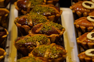 Rich chocolate tarts with golden pastry crusts topped with coffee beans and crushed pistachios are arranged neatly in metal trays, showcasing glossy textures and artisanal baking craftsmanship.


